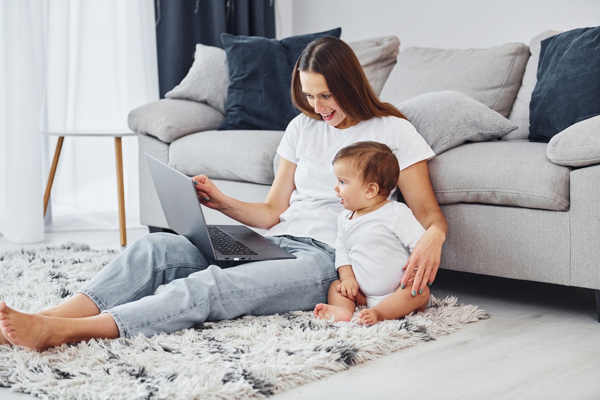 Silver colored laptop. Mother with her little daughter is indoors at home together